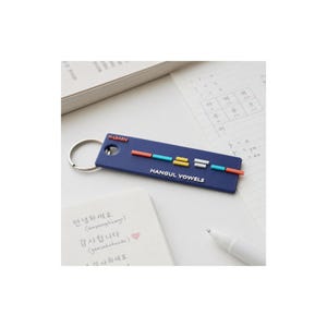 May include: A rectangular navy blue keychain featuring the text H-LEARN and HANGUL VOWELS in white. The center displays a series of small, colorful horizontal bars in orange, teal, yellow, and white, representing Korean vowel structures.