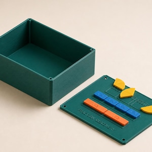 May include: A dark green rectangular storage box and a matching lid featuring the text "FRACTION DISCOVERY". The lid includes recessed slots containing orange, blue, and yellow geometric shapes representing fractions like 1/2 and 1/4.