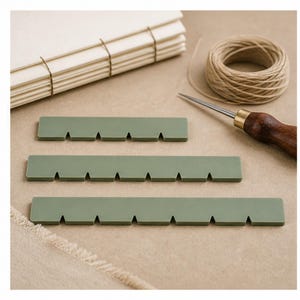 May include: Three sage green rectangular bookbinding alignment guides with V-shaped notches along one edge. These flat tools are arranged horizontally, accompanied by a spool of light tan thread, a metal awl with a wooden handle, and a stack of paper.