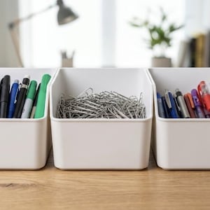 May include: Three white rectangular plastic storage bins arranged in a row. The left and right bins contain various colourful pens and markers, while the centre bin is filled with a large collection of silver metal paper clips.
