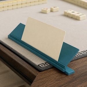 May include: A teal-colored card holder stand featuring a slanted back support and a front groove designed to hold a single rectangular card in an upright position. The stand is constructed with a smooth finish and a simple, functional design.