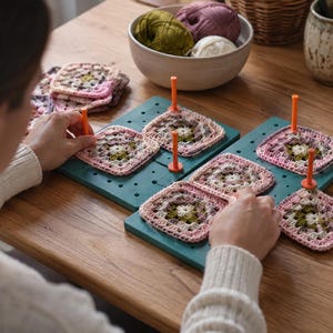 Op de afbeelding: Een vierkant teal-kleurig opspanbord met een raster van gaten en meerdere rechtopstaande oranje pinnen. Verschillende gehaakte granny squares in roze, groene en witte tinten zijn op de pinnen geplaatst om hun vorm tijdens het opspannen te behouden.
