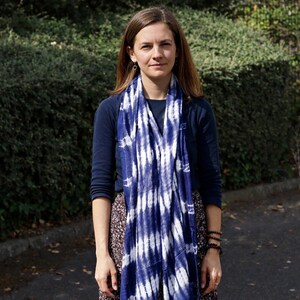 Peut inclure: Une longue écharpe rectangulaire présentant un motif tie-dye bleu profond et blanc avec des rayures verticales. L'écharpe est drapée autour du cou, tombant sur le devant d'un chemisier bleu foncé à manches longues et d'une jupe à motifs floraux.