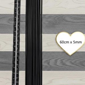 Może przedstawiać: Pęczek czarnych, smukłych prętów, prawdopodobnie do rękodzieła lub aranżacji kwiatowych, obok linijki. Grafika w kształcie serca wskazuje wymiary: 60cm x 5mm. Tło przedstawia wzór słojów drewna w odcieniach szarości i bieli.