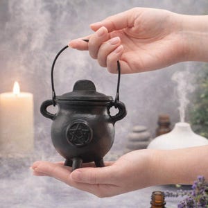 May include: A small, black cast iron cauldron with a lid and handle. The cauldron features a pentagram design on the front and is held by a person. The background includes a lit candle and a white diffuser, suggesting a mystical or spiritual theme.