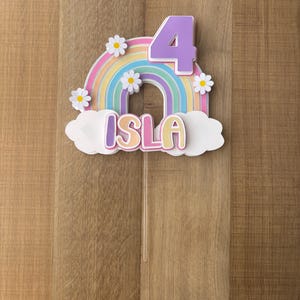 May include: A cake topper with a rainbow design, daisies, and the name "Isla". The number "4" is at the top. It is mounted on a clear stick. The background is a wooden surface. The measurements are in centimetres.