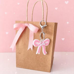 May include: A brown paper gift bag adorned with a pink ribbon. A pink bow-shaped keyring, inscribed with the name "Ayla", hangs from the bag. The background is pink, decorated with white hearts and sparkles.