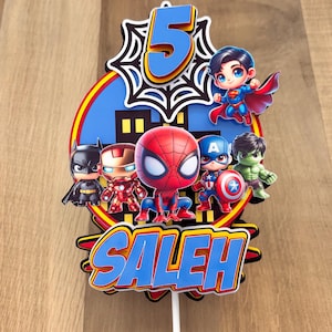 May include: Cake topper with superhero figures such as Spider-Man, Captain America, and Superman. The design includes the number 5 and the name "Saleh" in blue and yellow. The background is a blue circle with a cityscape.