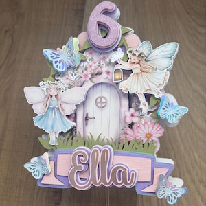 May include: A fairy-themed cake topper with the number 6, fairies, butterflies, flowers, and the name "Ella". The design uses pink, purple, blue, and green. The topper is designed to be inserted into a cake. The overall design is whimsical and decorative.