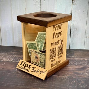 May include: A wooden tip jar with a dark brown top and base, and a light wood body. The jar has a slot on top and a small sign that reads "Tips Are Appreciated Thank you!" Inside are several US dollar bills. The side of the jar has the text "Your Logo Virtual Tip".