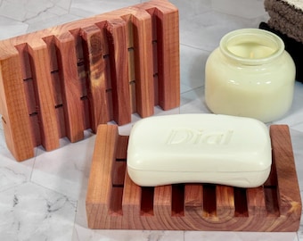 May include: Two wooden soap dishes made of reddish-brown cedar. One holds a bar of white soap with "Dial" printed on it. A jar candle and folded towels are in the background. The soap dishes have a slatted design.