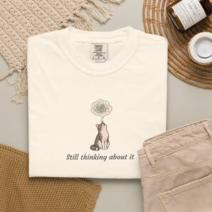 May include: A cream-colored short-sleeve t-shirt featuring a graphic of a brown and white tabby cat looking up at a thought bubble filled with scribbles. Below the cat, the text reads Still thinking about it in a dark brown serif font.
