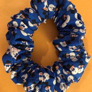 May include: A blue scrunchie with a snowman pattern. The scrunchie is made of fabric and has a gathered look.
