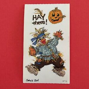 May include: A rectangular card with a cartoon scarecrow illustration. The scarecrow is wearing a blue plaid shirt, brown pants, and straw-filled boots. Above the scarecrow is a smiling jack-o'-lantern and the words "HAY there!" in black. The card is on a red background.