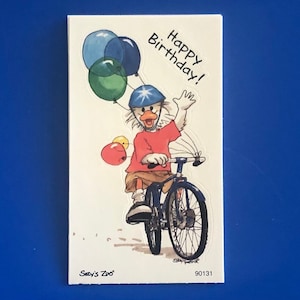 May include: A birthday card featuring a cartoon character riding a bicycle with balloons. The card says "Happy Birthday!"