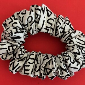 May include: A white scrunchie with a black, repeating abstract letter design. The fabric is gathered and elasticized, creating a textured, circular shape. The scrunchie is set against a solid red background.