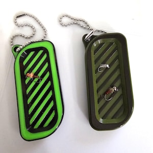 May include: Two rectangular fly fishing lure boxes with ball chain keychains. One box is green with black diagonal stripes and a fly. The other is olive green with diagonal stripes and two flies.