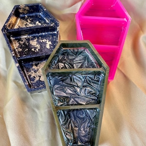 Peut inclure: Trois plateaux en forme de cercueil de couleurs variées. L'un est bleu marine avec des accents de feuilles d'or, un autre rose vif et le troisième vert olive avec un intérieur texturé et métallique. Les plateaux sont probablement destinés à un usage décoratif ou organisationnel.