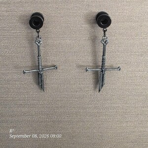 May include: A pair of black ear plugs with dangling silver-toned sword pendants. Each earring features a detailed hilt and a pointed blade design hanging from a circular black plug. The metal has a weathered, dark finish with visible texture.