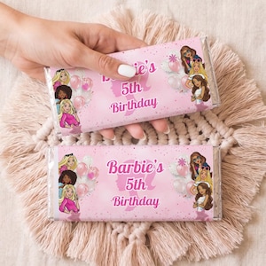 Puede incluir: Dos envoltorios rosas de barras de caramelo con el texto "Barbie's 5th Birthday" e ilustraciones de muñecas Barbie y globos. Los envoltorios están sostenidos por una mano, descansando sobre un tapete beige tejido.