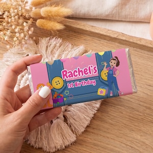 May include: A pink and blue candy bar wrapper with the text "Rachel's 1st Birthday" and a cartoon illustration of a girl in overalls. The wrapper has a denim design with yellow buttons and stars.