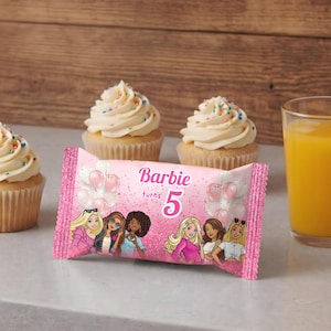 May include: A festive scene with cupcakes, a juice glass, and a pink candy wrapper. The cupcakes have white frosting and colorful sprinkles. The wrapper says "Barbie turns 5" and features Barbie characters. A glass of orange juice with an orange slice.