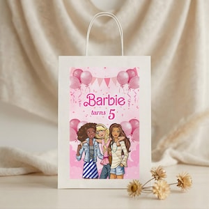 May include: White paper gift bag with a pink graphic featuring the text "Barbie turns 5" and illustrations of three girls and pink balloons. The bag has a white handle.