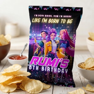 Op de afbeelding: Een zwarte zak chips met de tekst "I'M DONE HIDING, NOW I'M SHINING LIKE I'M BORN TO BE" en "RUMI'S 8TH BIRTHDAY". De zak heeft een afbeelding van drie geanimeerde vrouwen. Schalen met chips en een kleine kom ketchup staan op de achtergrond.