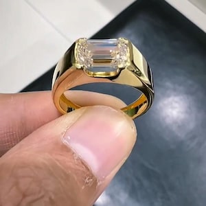 Op de afbeelding: Een gouden ring met een rechthoekige, heldere edelsteen en kleinere accentstenen. De ring heeft een dikke band en een gepolijste afwerking, die een klassiek ontwerp laat zien. De ring wordt vastgehouden door de vingers van een persoon.