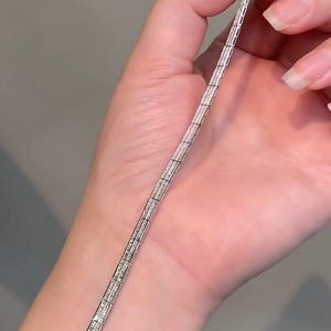 Pu&ograve; includere: Un bracciale in metallo color argento caratterizzato da una serie di maglie rettangolari, ciascuna incastonata con molteplici piccole pietre chiare e scintillanti disposte a griglia. Il bracciale presenta una chiusura sicura e finitura lucida.