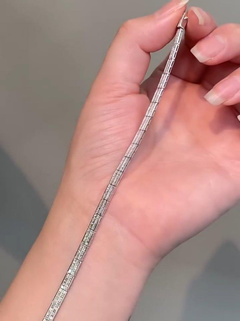 Pu&ograve; includere: Un bracciale in metallo color argento caratterizzato da una serie di maglie rettangolari, ciascuna incastonata con molteplici piccole pietre chiare e scintillanti disposte a griglia. Il bracciale presenta una chiusura sicura e finitura lucida.