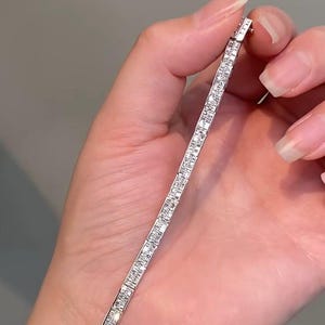 Pu&ograve; includere: Un bracciale tennis color argento caratterizzato da una fila continua di piccole pietre trasparenti a taglio rettangolare incastonate in un canale metallico. Il bracciale ha un design lineare e sottile con una finitura lucida che riflette la luce.