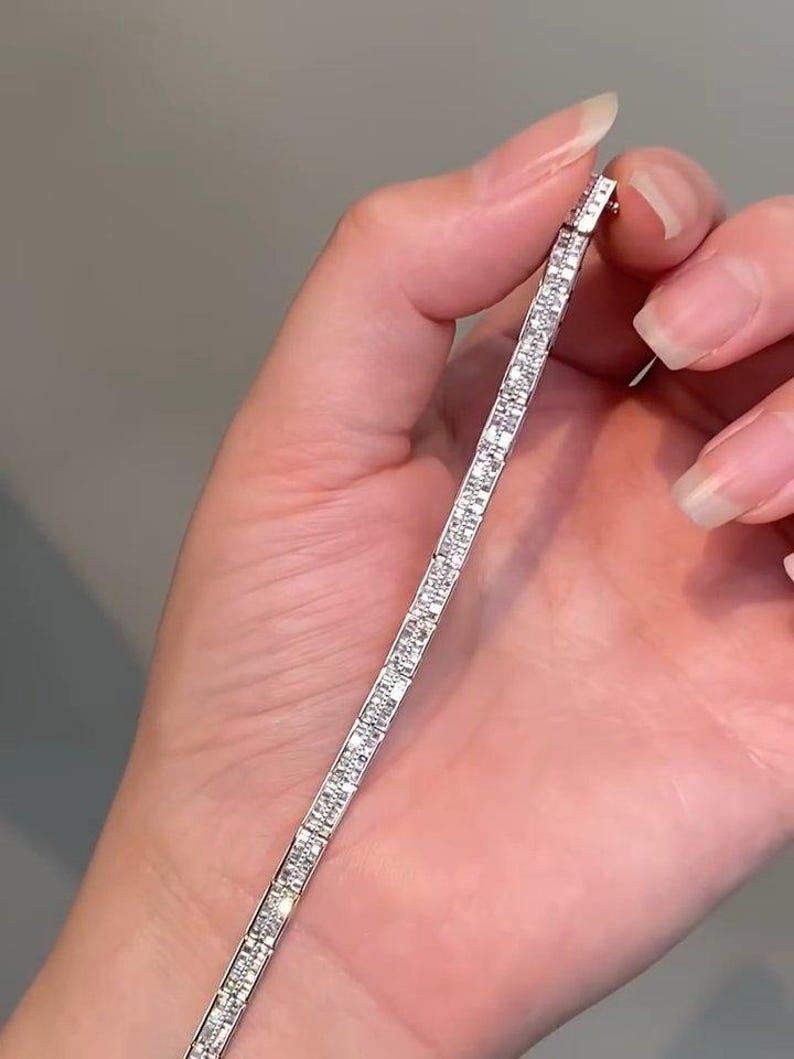 Pu&ograve; includere: Un bracciale tennis color argento caratterizzato da una fila continua di piccole pietre trasparenti a taglio rettangolare incastonate in un canale metallico. Il bracciale ha un design lineare e sottile con una finitura lucida che riflette la luce.