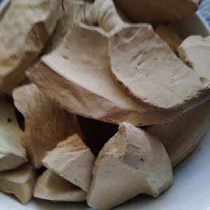 May include: A white bowl containing several irregularly shaped, light brown pieces. The pieces have a rough, textured surface and vary in size. The lighting creates shadows, adding depth to the image. The bowl is plain and white.