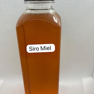 May include: A clear plastic bottle filled with amber liquid and a black cap. The label on the bottle reads "Siro Miel". The bottle has a rectangular shape with rounded corners.