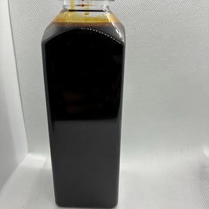 May include: A clear, square-shaped plastic bottle filled with a dark brown liquid. The bottle features a black, ribbed screw-top cap. The liquid inside is opaque and dark, filling the container nearly to the top, with a small amount of residue visible.