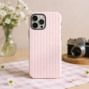 May include: A pink and white striped phone case. The case is on a wooden table with a vintage camera and a vase of white flowers in the background. The case has a black camera lens.