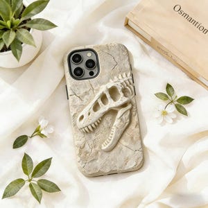 May include: A beige phone case featuring a 3D dinosaur skull design. The case has a textured, cracked appearance, and the skull is a lighter beige color. The phone case is surrounded by white fabric, small white flowers, and greenery.