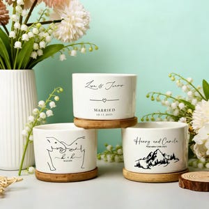 以下が含まれることがあります： 木製のベースが付いた3つの白いセラミックポット。1つのポットには「Lisa & Travis」という名前と日付付きの「MARRIED」という言葉が書かれています。別のポットには象のイラストとイニシャルが描かれています。3番目のポットには名前と山の風景があります。