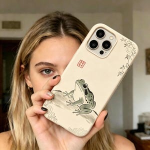 May include: A cream-colored phone case featuring a detailed illustration of a frog. The frog is depicted in shades of gray and green, with a black eye and a speckled pattern. The case also has a small red square with Japanese characters.