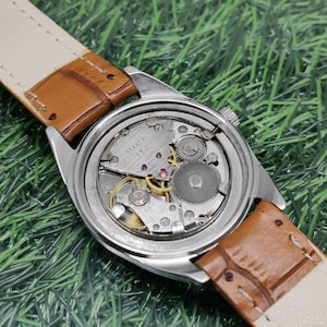 May include: A vintage wristwatch with a silver-colored case and a brown leather strap. The watch face is visible, showcasing the intricate internal mechanisms. The strap is a light tan color on the inside. The watch is set against a green background.