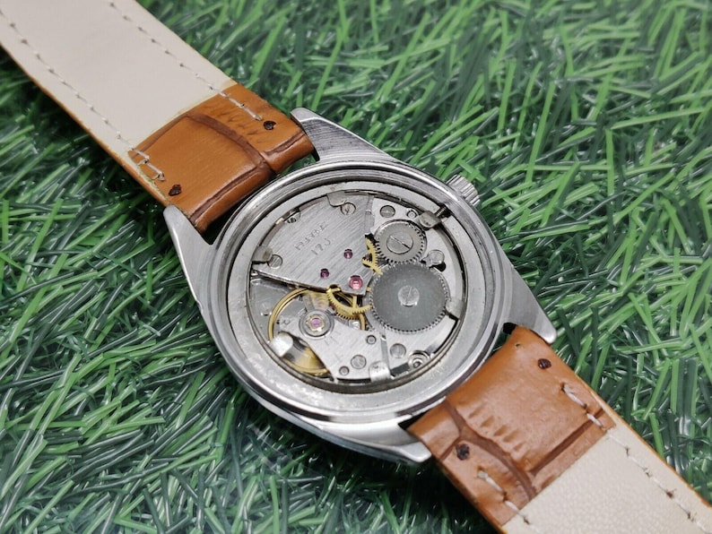 May include: A vintage wristwatch with a silver-colored case and a brown leather strap. The watch face is visible, showcasing the intricate internal mechanisms. The strap is a light tan color on the inside. The watch is set against a green background.