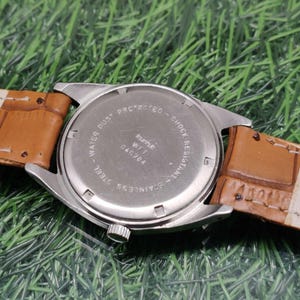 May include: Close-up of the back of a wristwatch with a stainless steel case and a brown leather strap. The watch face is silver with the text "WATER DUST PROTECTED - SHOCK RESISTANT - STAINLESS STEEL". The strap is a light brown color.