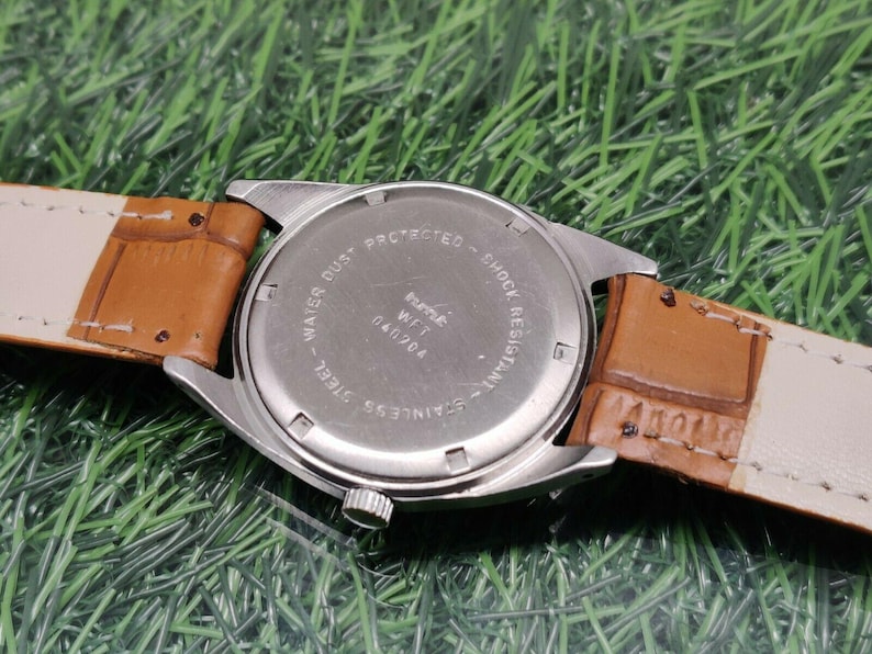 May include: Close-up of the back of a wristwatch with a stainless steel case and a brown leather strap. The watch face is silver with the text "WATER DUST PROTECTED - SHOCK RESISTANT - STAINLESS STEEL". The strap is a light brown color.