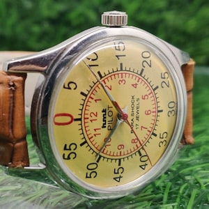 May include: A vintage wristwatch with a silver case and a yellow dial. The dial features black numerals, red accents, and the text "hmt PILOT". The watch has a brown leather strap.
