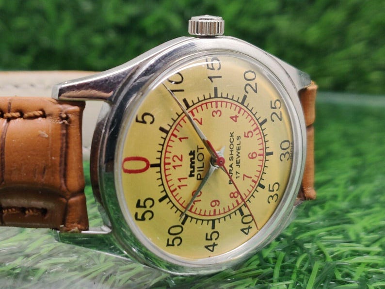 May include: A vintage wristwatch with a silver case and a yellow dial. The dial features black numerals, red accents, and the text "hmt PILOT". The watch has a brown leather strap.