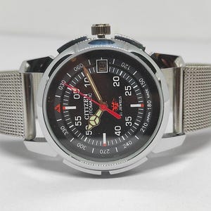 Op de afbeelding: Een Citizen automatisch polshorloge met een zwarte wijzerplaat, zilverkleurige kast en mesh band. De wijzerplaat heeft witte en rode markeringen, waaronder "Citizen Automatic 21 Jewels". Datumweergave op 3 uur.