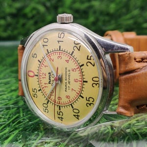 May include: A vintage wristwatch with a silver case and a light yellow face. The watch features a red inner dial with numbers and the text "PARA SHOCK 17 JEWELS". The watch has a brown leather strap.