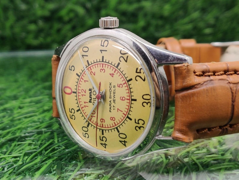 May include: A vintage wristwatch with a silver case and a light yellow face. The watch features a red inner dial with numbers and the text "PARA SHOCK 17 JEWELS". The watch has a brown leather strap.