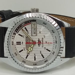 Pode incluir: Um relógio automático Citizen prateado com uma bracelete de couro preta. O mostrador branco apresenta detalhes em preto e vermelho, mostrando o dia e a data. O relógio tem uma moldura texturizada e uma bracelete de couro preta.